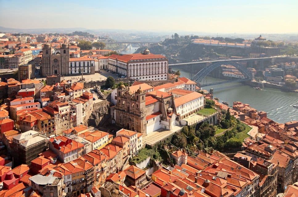 A high-angle view of a historic city with red-tiled roofs, a large river, and an arched bridge on a sunny day.