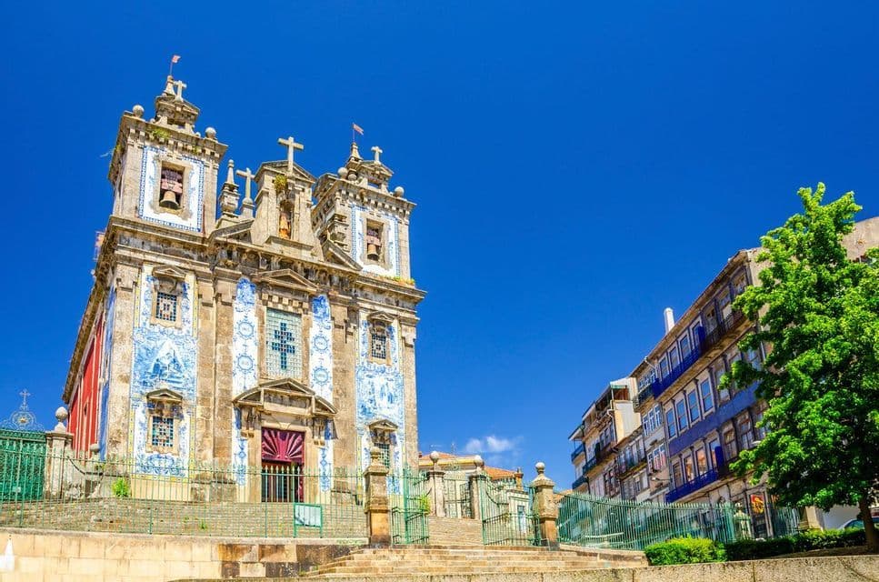 An ornate church with a blue and white tiled facade stands on a stone terrace under a clear blue sky.