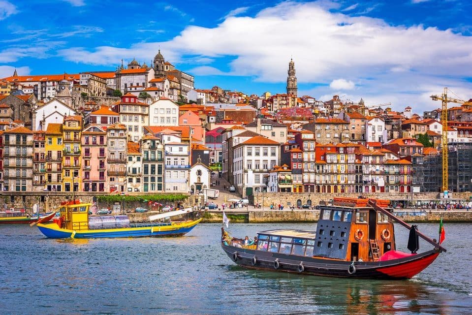 Two traditional boats sail on a river in front of a colorful hillside city with a prominent stone tower under a blue sky.