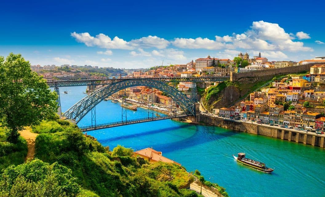 A large metal arch bridge spans a blue river next to a hillside city with colorful buildings, with a tour boat sailing below.