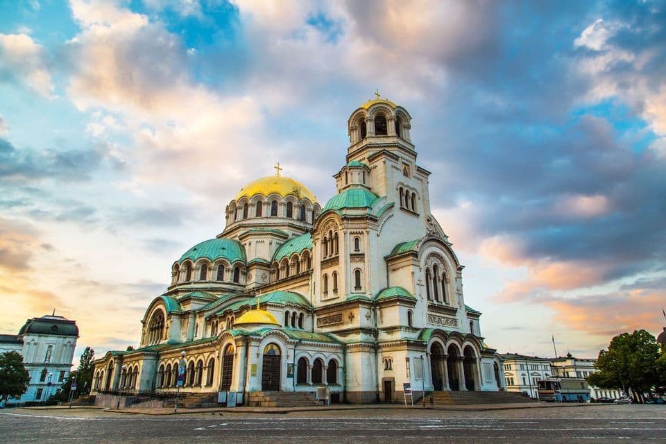 A large white cathedral with golden and green domes stands in a square under a cloudy sky at sunset.