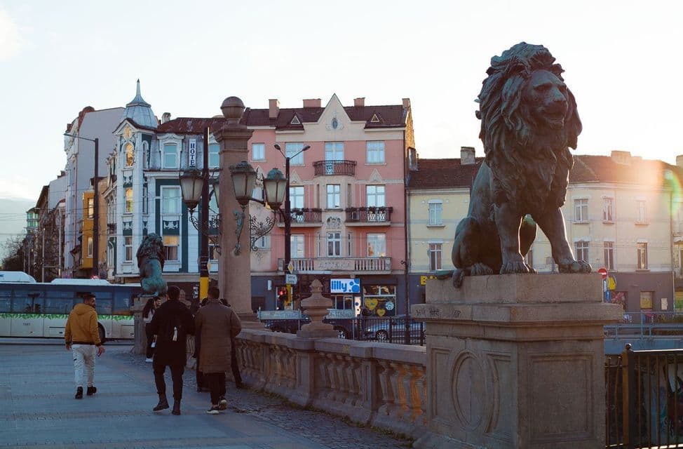 A WeRoad group trip walks on a bridge next to a large bronze lion statue, with colorful city buildings in the background.