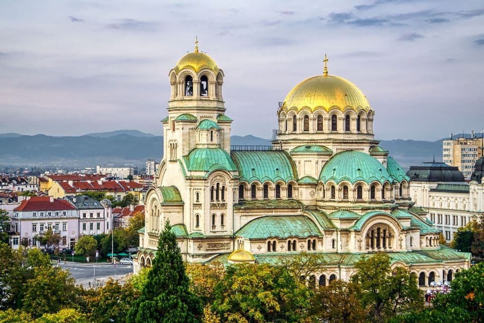 A large cathedral with golden and green domes overlooks a cityscape with trees in the foreground and mountains in the background.