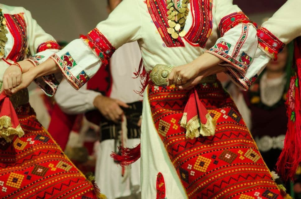 A close-up of dancers holding hands, wearing traditional costumes with red embroidered patterns and metal accessories.