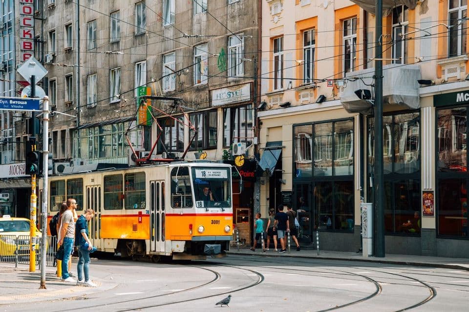 A yellow and white tram travels along tracks on a city street, with people standing on the sidewalk next to tall buildings.