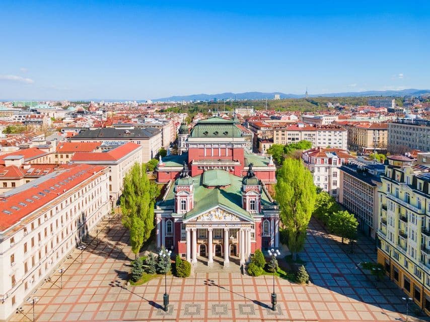 An aerial view of a grand, ornate red theater with a green roof, situated in a large plaza surrounded by a cityscape under a clear blue sky.