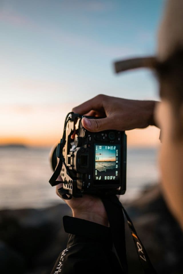Ein Blick über die Schulter einer Person, die eine Digitalkamera hält und das Meer während des Sonnenuntergangs fotografiert.