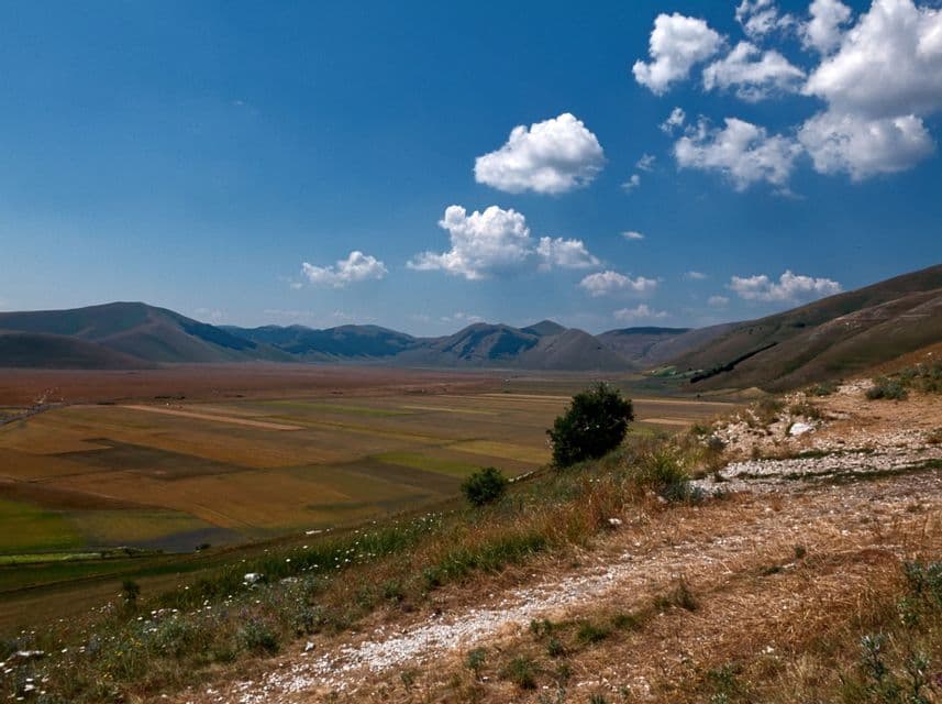 Una vasta valle di campi a mosaico e montagne ondulate sotto un cielo azzurro brillante con nuvole bianche, vista da un pendio erboso.