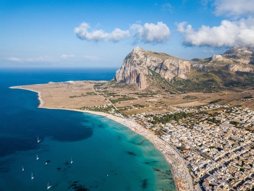 Vista aérea de un pueblo costero con una playa concurrida junto a agua turquesa, al lado de una gran montaña rocosa bajo un cielo azul.