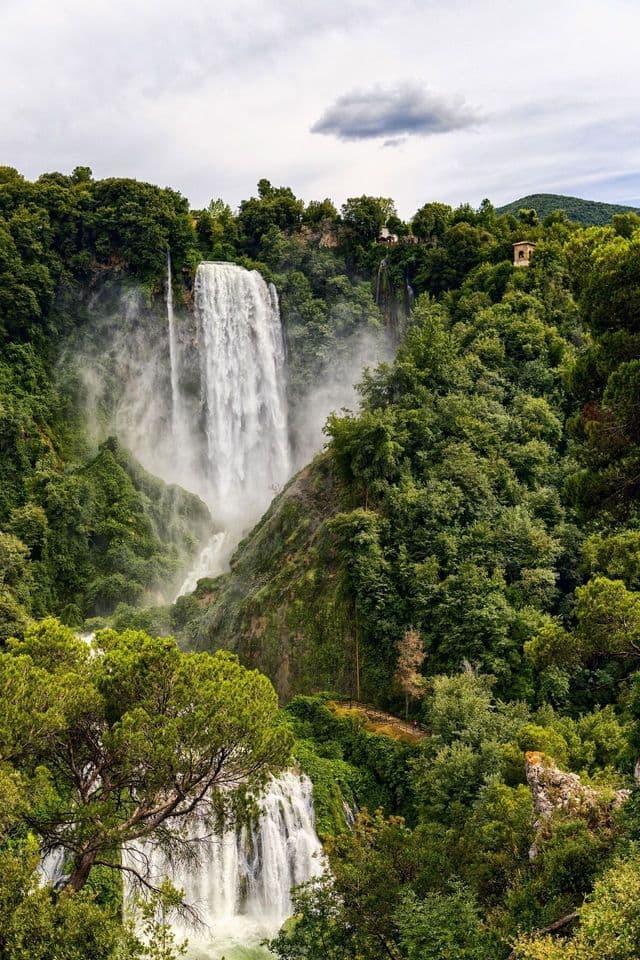 Una cascata a più livelli scende da una scogliera circondata da una fitta foresta verde sotto un cielo parzialmente nuvoloso.