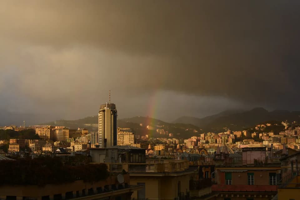 A cityscape with a tall skyscraper under a dark, stormy sky, with a rainbow arching over distant hills.
