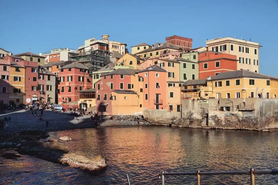 Colorful buildings on a hillside overlook a small pebble beach where people relax by the water under a clear blue sky.