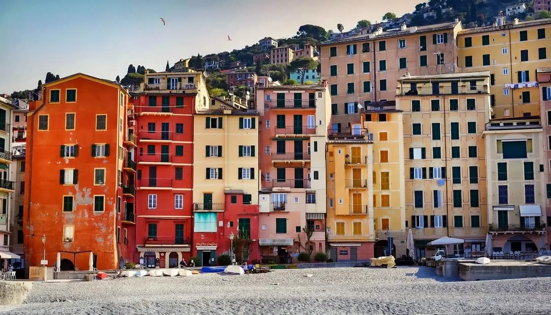 A row of colorful, multi-story buildings stands on a pebble beach, with a green hillside rising in the background under a clear sky.