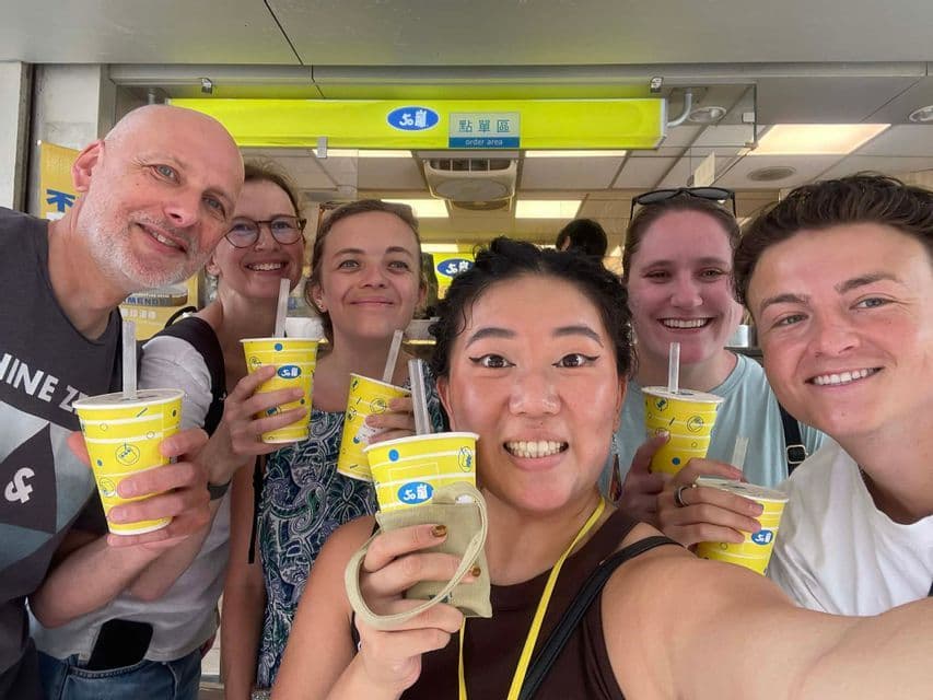 Un voyage de groupe WeRoad sourit pour un selfie, tenant des gobelets jaunes avec des pailles devant un comptoir de magasin.