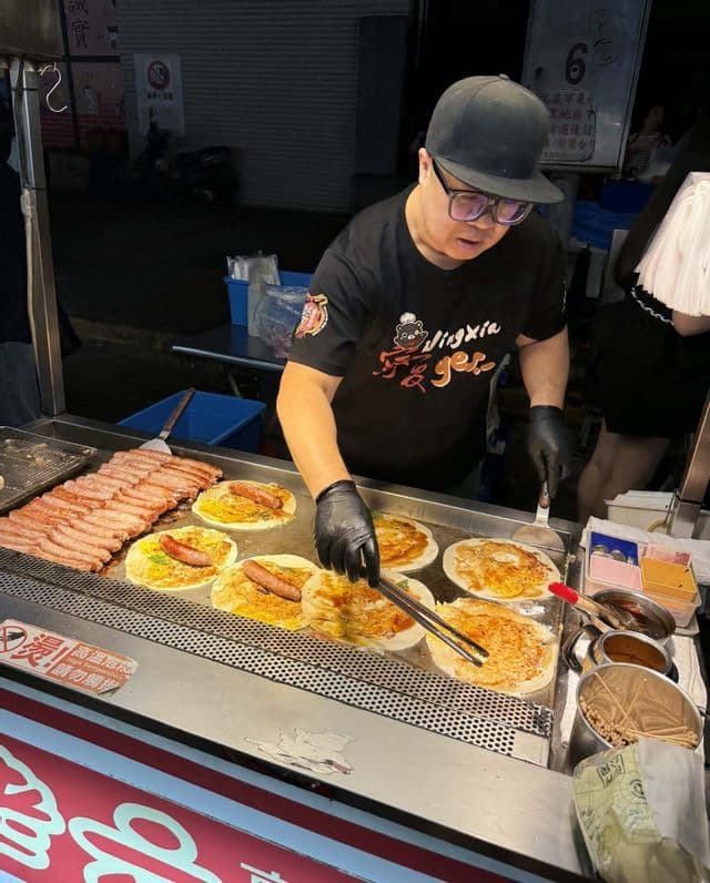 Un vendeur de street food, coiffé d'une casquette et ganté, cuisine des wraps saucisse-œuf sur une grande plancha, sur un marché de nuit.