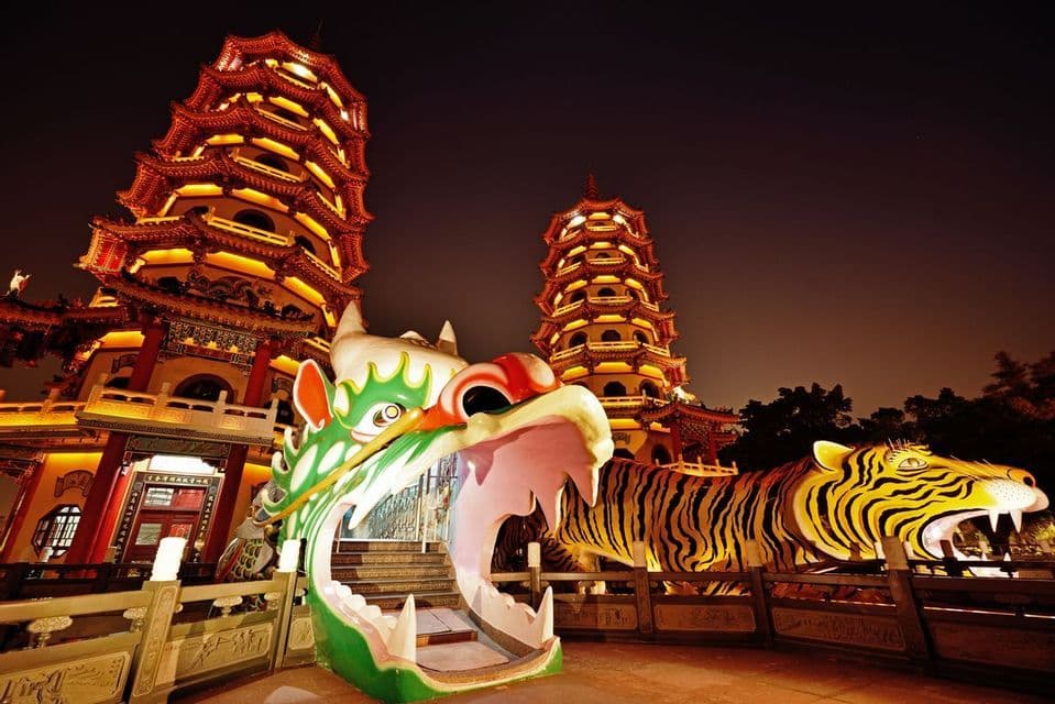 De grandes statues d'un dragon et d'un tigre se dressent devant deux pagodes illuminées la nuit.