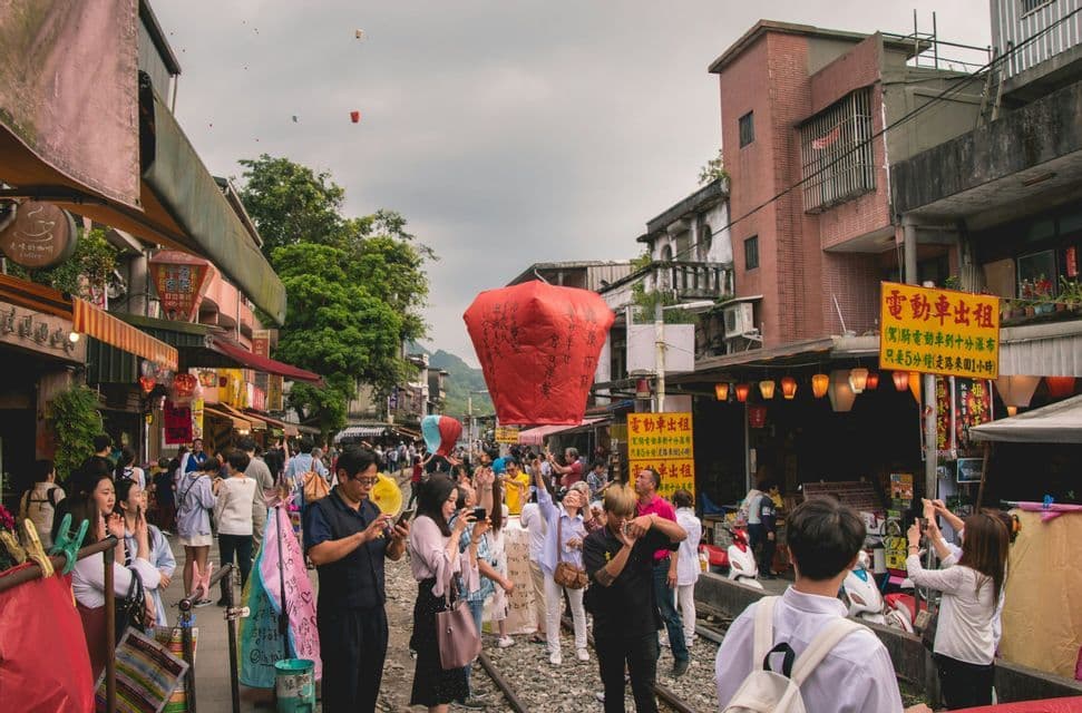 Une foule de personnes sur des voies ferrées, dans une rue animée, lâche une grande lanterne en papier rouge dans le ciel nuageux.