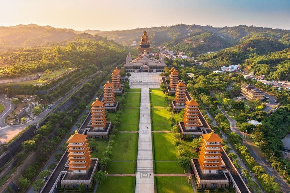 Une vue aérienne d'un grand complexe de temples où huit pagodes bordent un chemin menant à une statue géante de Bouddha assis sur une colline verdoyante et luxuriante.