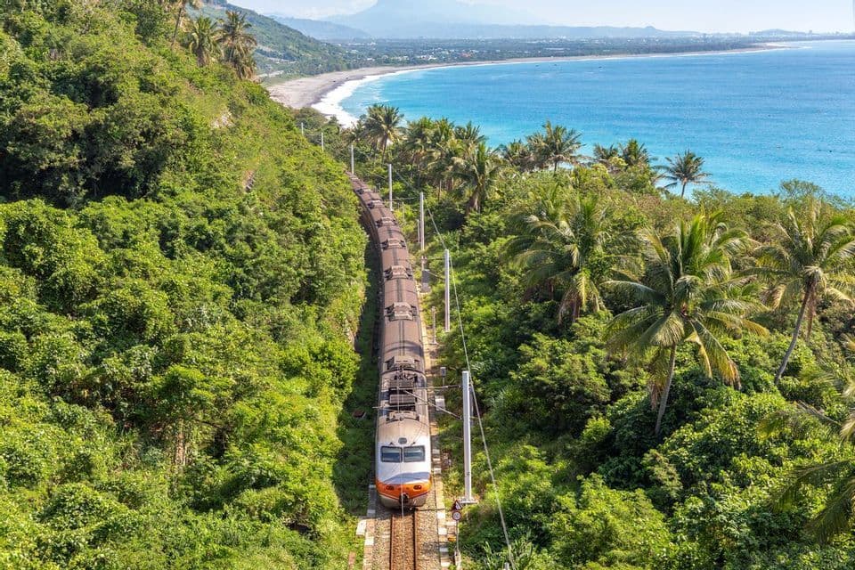 Vue aérienne d'un train circulant sur des rails à travers une forêt verdoyante et luxuriante, le long d'un littoral sablonneux et de l'océan.