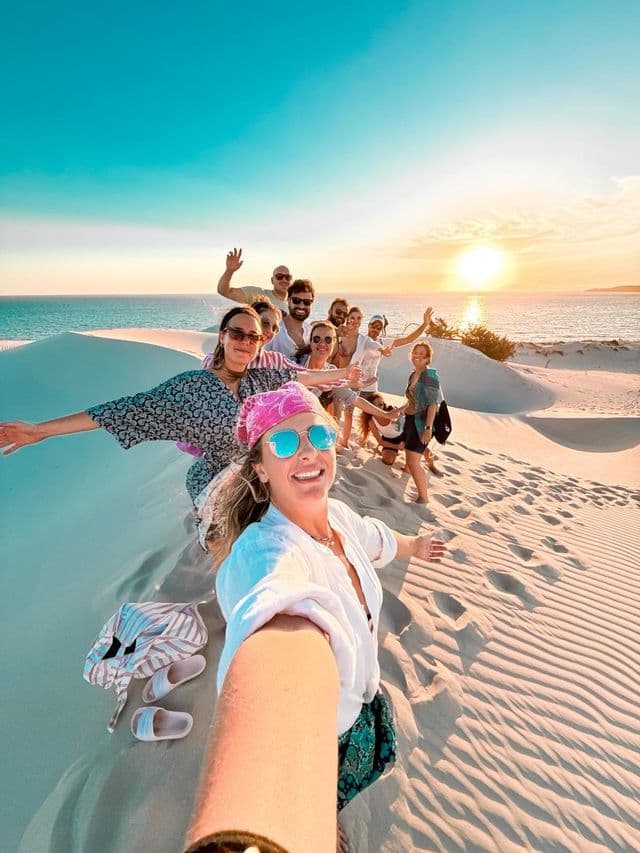 Un viaggio di gruppo WeRoad scatta un selfie sorridente sulle dune di sabbia bianca mentre il sole tramonta sull'oceano.