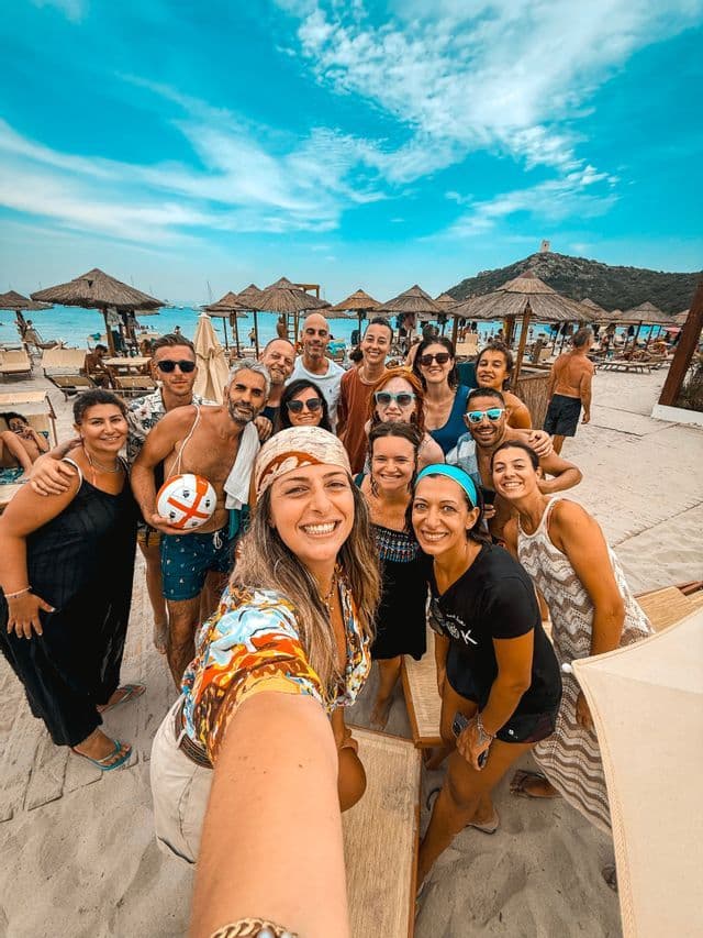 Un gruppo WeRoad si scatta un selfie sorridente su una spiaggia sabbiosa e affollata con ombrelloni di paglia sotto un cielo azzurro.