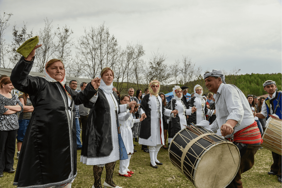 Un gruppo di persone in costumi tradizionali balla in fila, tenendosi per mano, accompagnato da un uomo che suona un grande tamburo all'aperto.