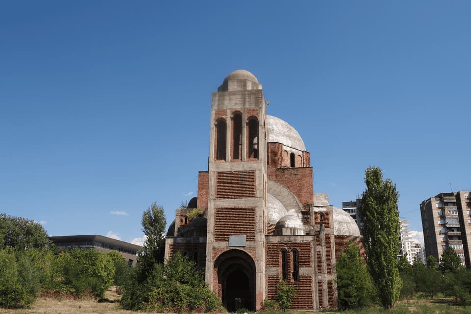Una chiesa incompiuta in mattoni rossi e cemento con cupole multiple si erge in un campo erboso sotto un cielo azzurro e limpido.