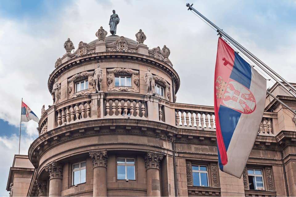 Un sontuoso edificio classico con tetto a cupola e statue, e due bandiere serbe che sventolano contro un cielo nuvoloso.