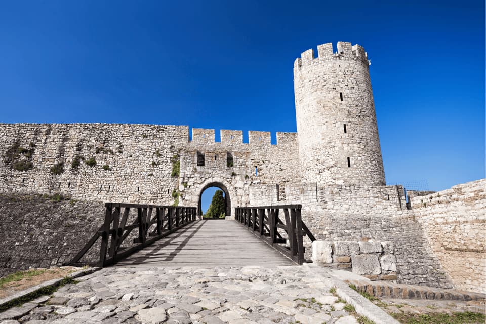 Un ponte di legno su un sentiero acciottolato conduce all'ingresso ad arco di una fortezza in pietra con una torre rotonda, sotto un cielo azzurro splendente.