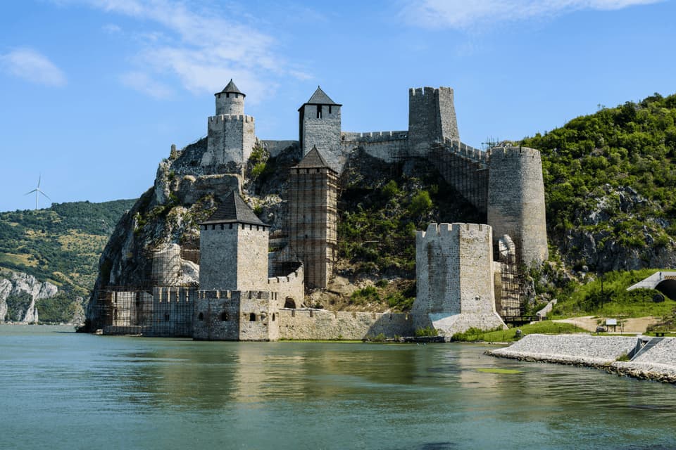 Una fortezza di pietra con più torri si erge su una collina rocciosa accanto a un ampio fiume sotto un cielo parzialmente nuvoloso.