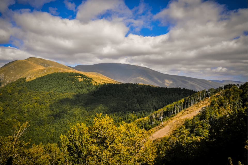 Un'ampia veduta di una catena montuosa con lussureggianti foreste verdi e colline dorate, con una seggiovia che sale su un pendio sotto un cielo nuvoloso.