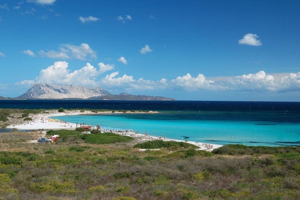 Vista elevata di una spiaggia di sabbia bianca accanto ad acqua turchese, con una grande isola rocciosa in lontananza sotto un cielo blu e nuvoloso.