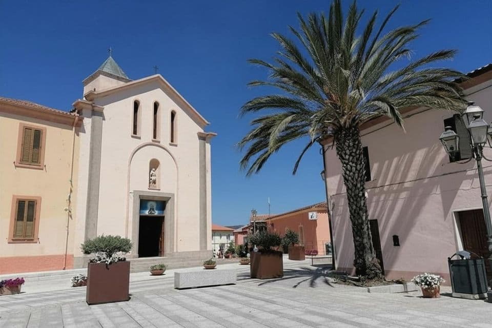 Una chiesa rosa pallido si erge su una piazza pavimentata accanto a una grande palma sotto un cielo azzurro limpido.
