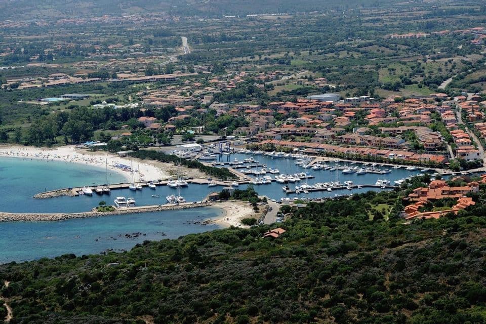 Una vista aerea di un porto turistico costiero pieno di barche, accanto a una spiaggia sabbiosa e una città con case dai tetti rossi.