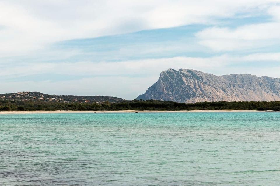Panoramica su acqua turchese, spiaggia di sabbia e imponente montagna rocciosa sotto un cielo parzialmente nuvoloso.