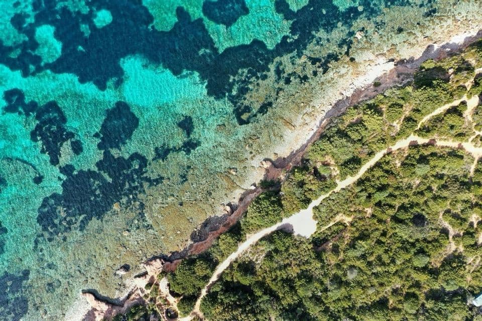 Veduta aerea di un sentiero costiero tortuoso che costeggia una riva rocciosa con acqua limpida e turchese.