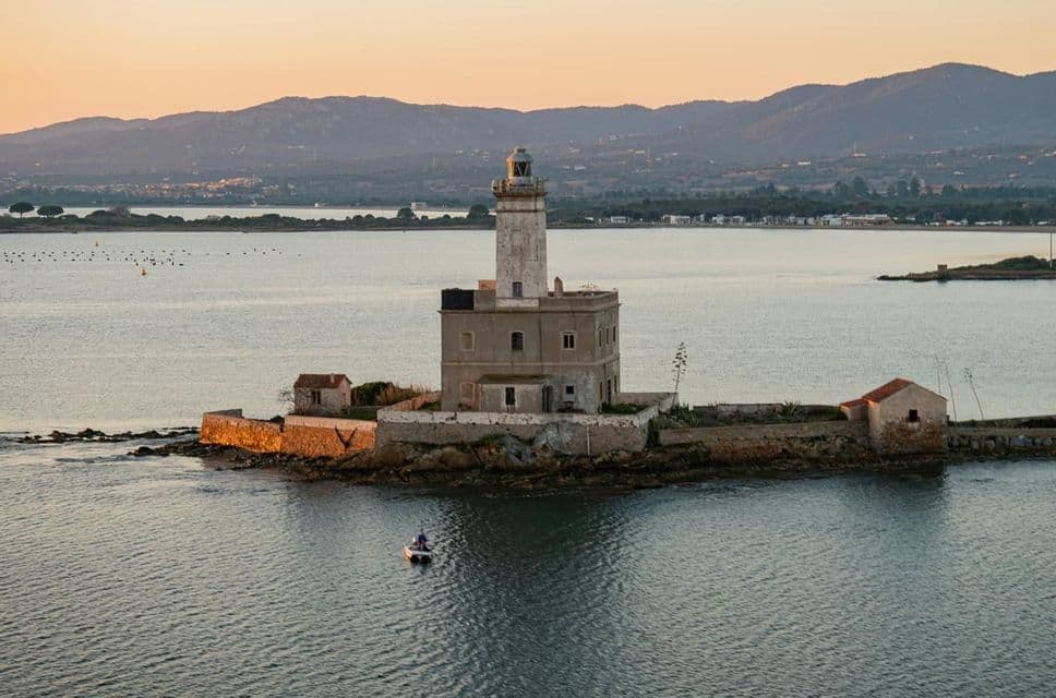 Un faro in pietra su una piccola isola rocciosa al tramonto, con una barca sull'acqua e montagne in lontananza.