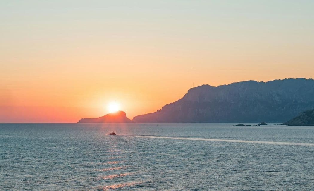 Una piccola barca naviga sul mare calmo mentre il sole tramonta dietro una costa rocciosa in silhouette, proiettando un bagliore dorato.