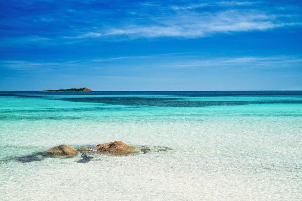 Rocce affiorano in acque basse, limpide e turchesi, con un'isola distante all'orizzonte sotto un cielo azzurro.