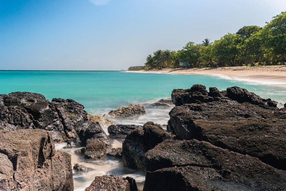 Lunga esposizione di acqua turchese che scorre tra scure rocce vulcaniche su una spiaggia sabbiosa fiancheggiata da palme sotto un cielo azzurro.