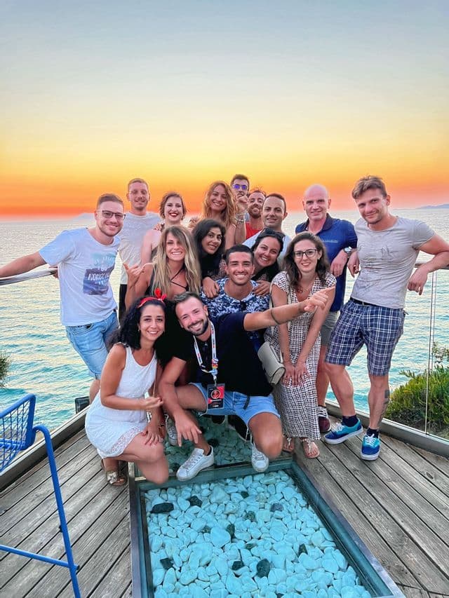 Un gruppo WeRoad posa per una foto su una terrazza in legno con pavimento in vetro, affacciata sul mare al tramonto.