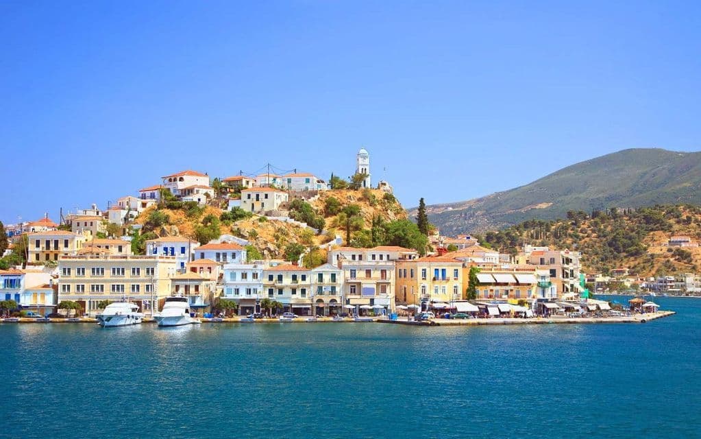 Una cittadina costiera con case colorate e una torre dell'orologio si erge su una collina sul mare azzurro brillante, con barche ormeggiate nel porto.