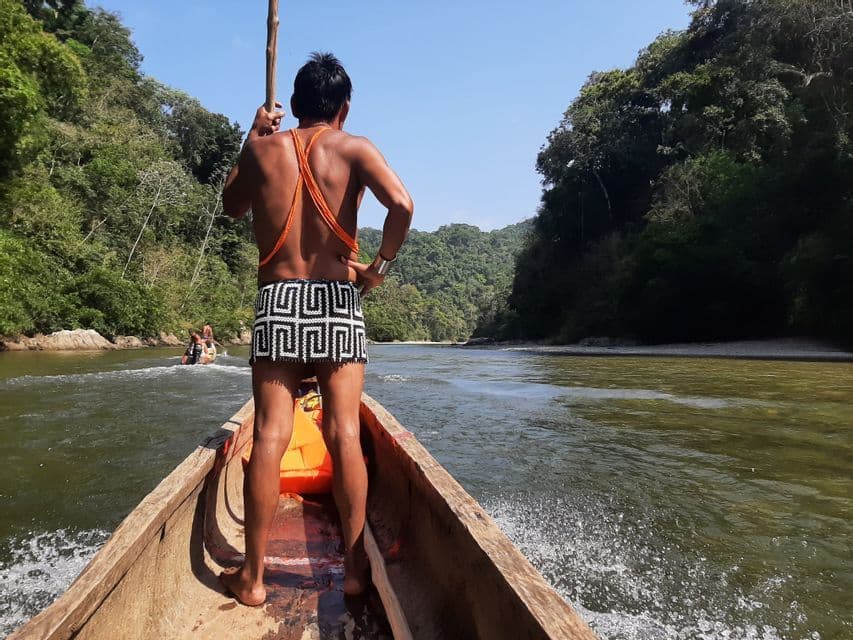 Un homme en tenue traditionnelle se tient à la proue d'un canoë en bois, le dirigeant avec une perche le long d'une rivière bordée d'une forêt luxuriante.