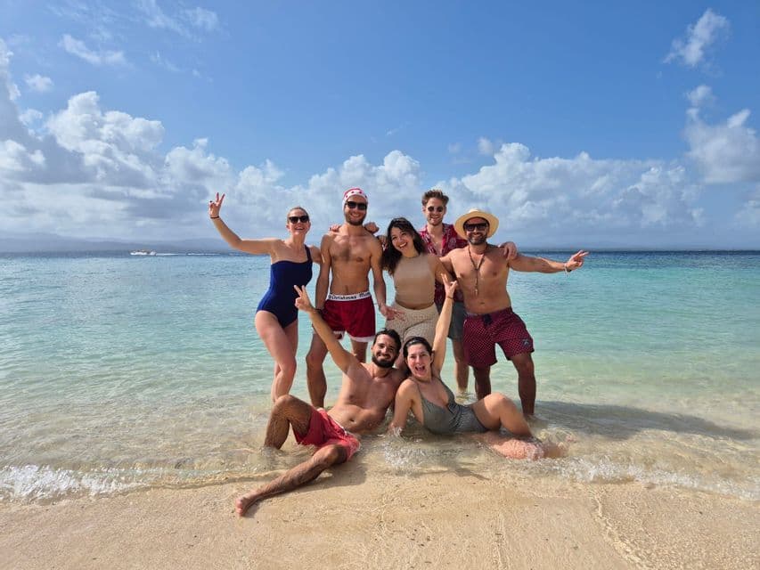 Un voyage de groupe WeRoad de sept personnes en maillot de bain posant pour une photo dans l'eau peu profonde et claire d'une plage de sable.