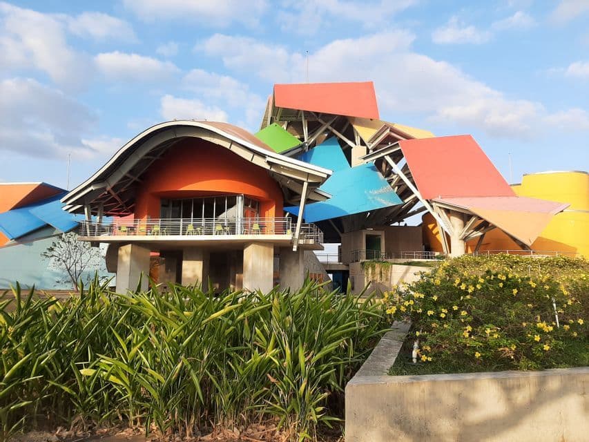 Un bâtiment moderne coloré avec des toits abstraits et superposés se dresse derrière des plantes vertes luxuriantes sous un ciel partiellement nuageux.