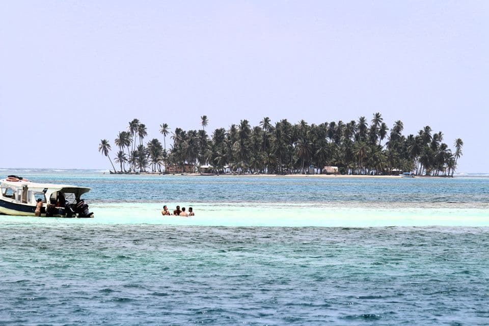 Un groupe WeRoad en voyage, nageant dans des eaux turquoises peu profondes près d'un bateau, avec une île tropicale bordée de palmiers en arrière-plan.