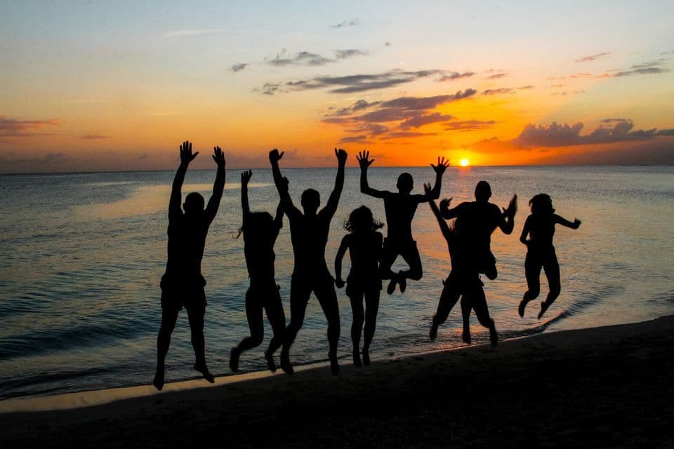 Silhouettes d'un groupe WeRoad sautant sur une plage de sable au coucher du soleil sur l'océan.