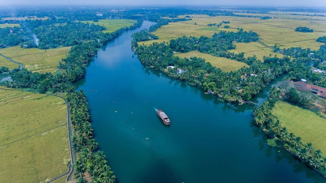 Una vista aérea de una casa flotante navegando por un ancho río azul, rodeada de exuberantes palmeras verdes y campos agrícolas.