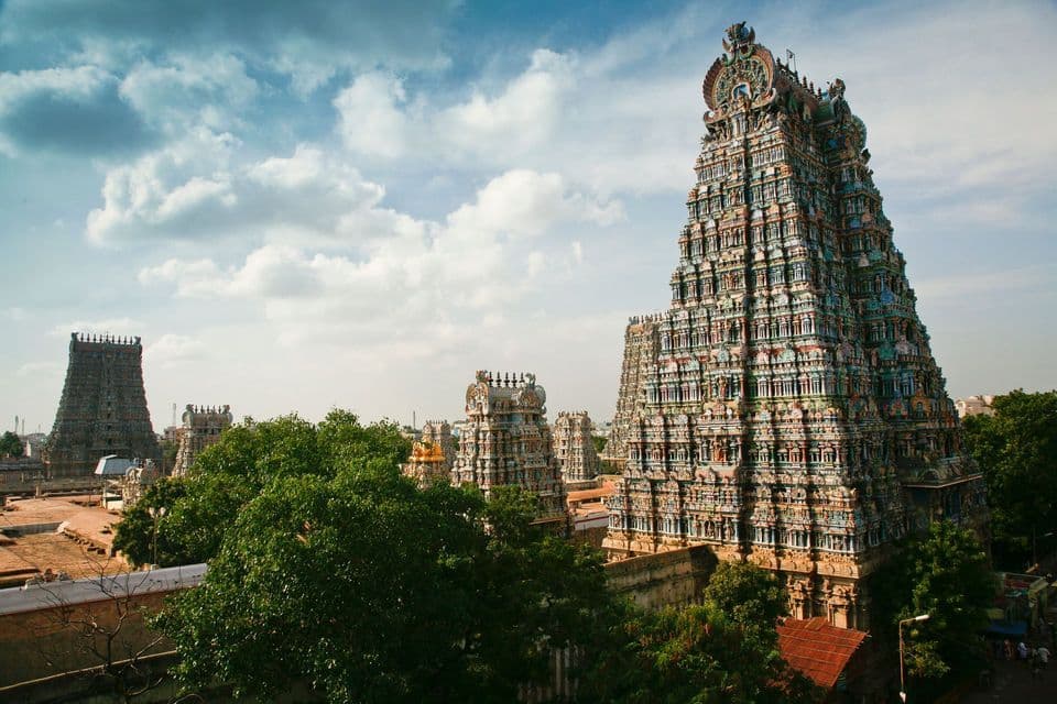 Les tours de temples hindous ornées et colorées s'élèvent au-dessus des cimes verdoyantes des arbres sous un ciel bleu partiellement nuageux.