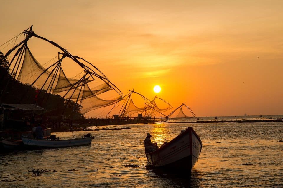 Una piccola barca con una persona sull'acqua, vicino a reti da pesca cinesi stagliate contro un tramonto dorato.